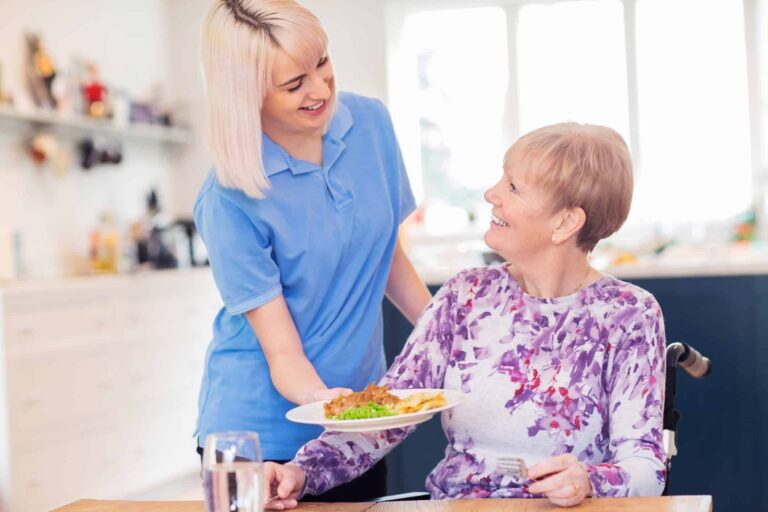 Female Care Assistant Serving Meal To Senior Woman Seated In Wheelchair At Table